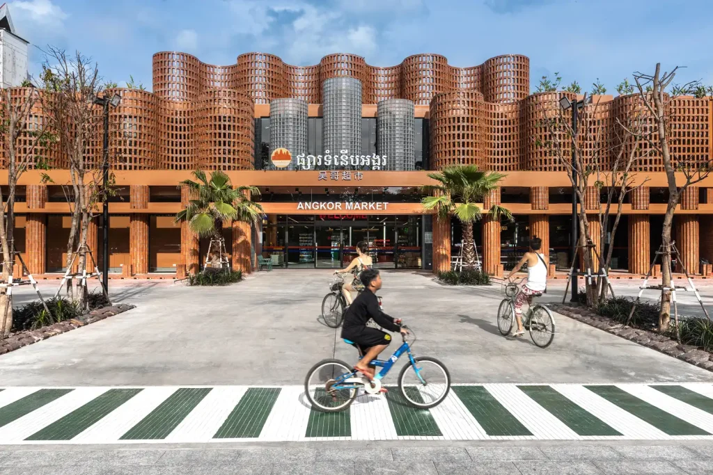 The front view of Angkor Market, designed by UAD Architects, featuring a wavy brick façade with glass cylinders and cyclists passing by in the foreground